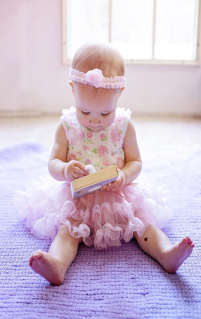 A young toddler girl in a pink frilly dress examining a book, seated on the floor.